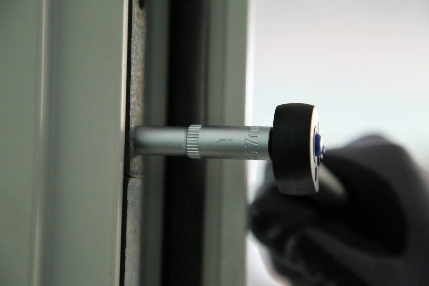 Dłoń w czarnej rękawiczce obraca narzędzie przy framudze okiennej, prawdopodobnie podczas prac montażowych lub czyszczących.