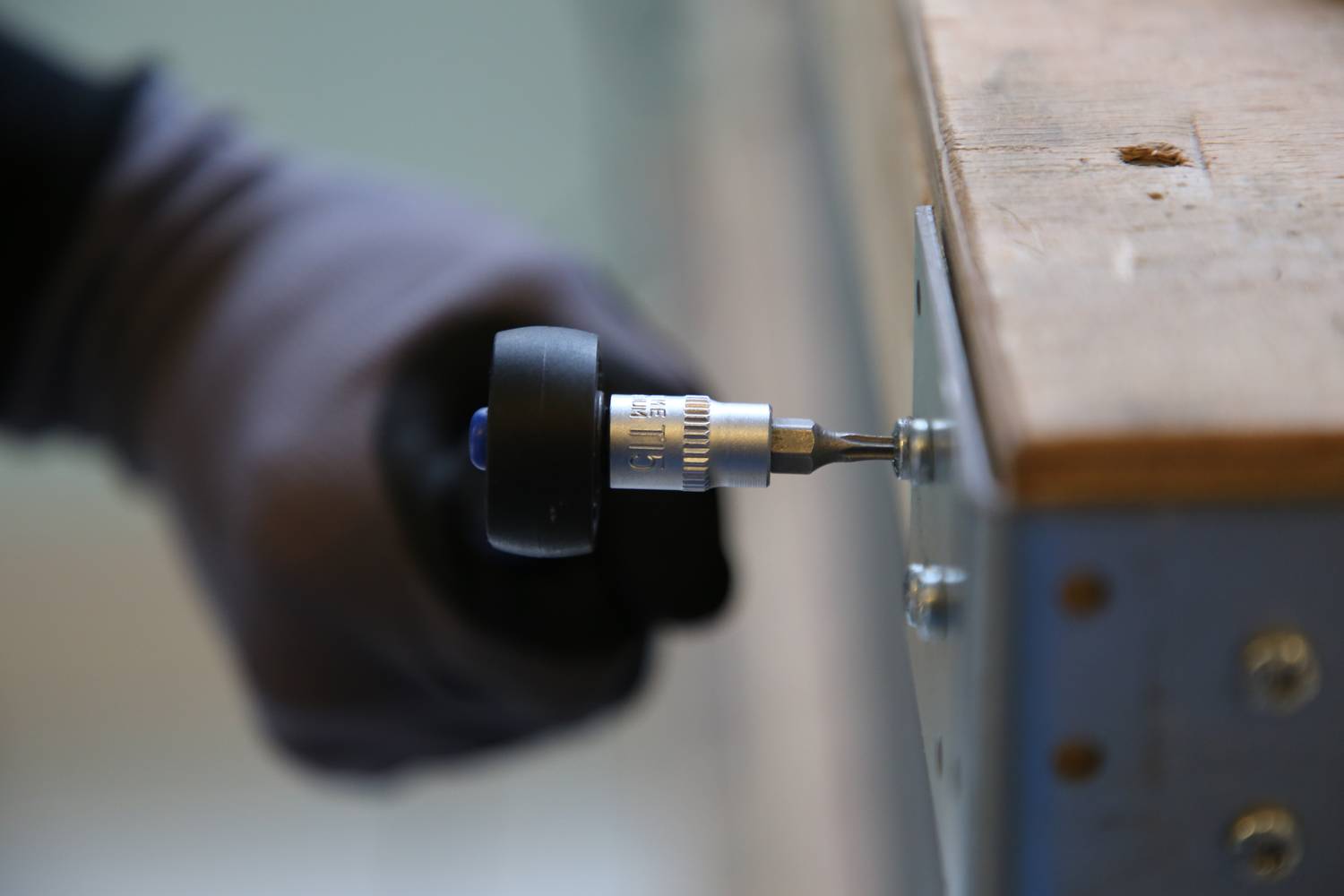 Osoba dokręca wkręt akumulatorowym wkrętakiem przy drewnianym stole. Fokus jest skierowany na wkręt i narzędzie.