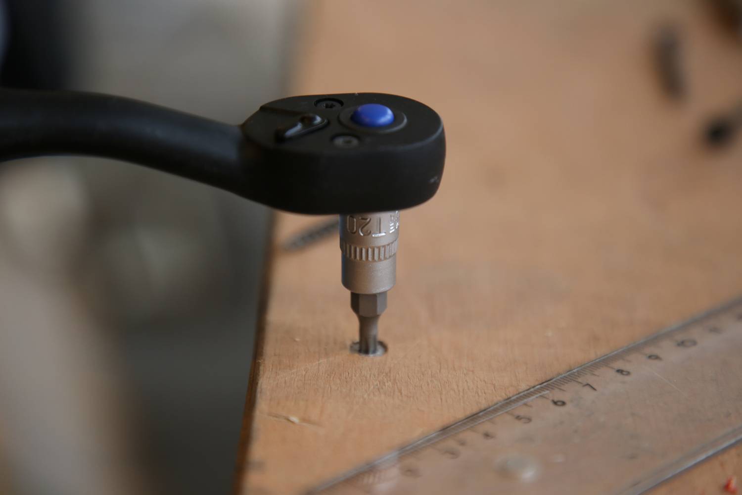 Akumulatorowa wkrętarka wkręca śrubę w drewnianą płytę. Obok leży metalowa linijka.