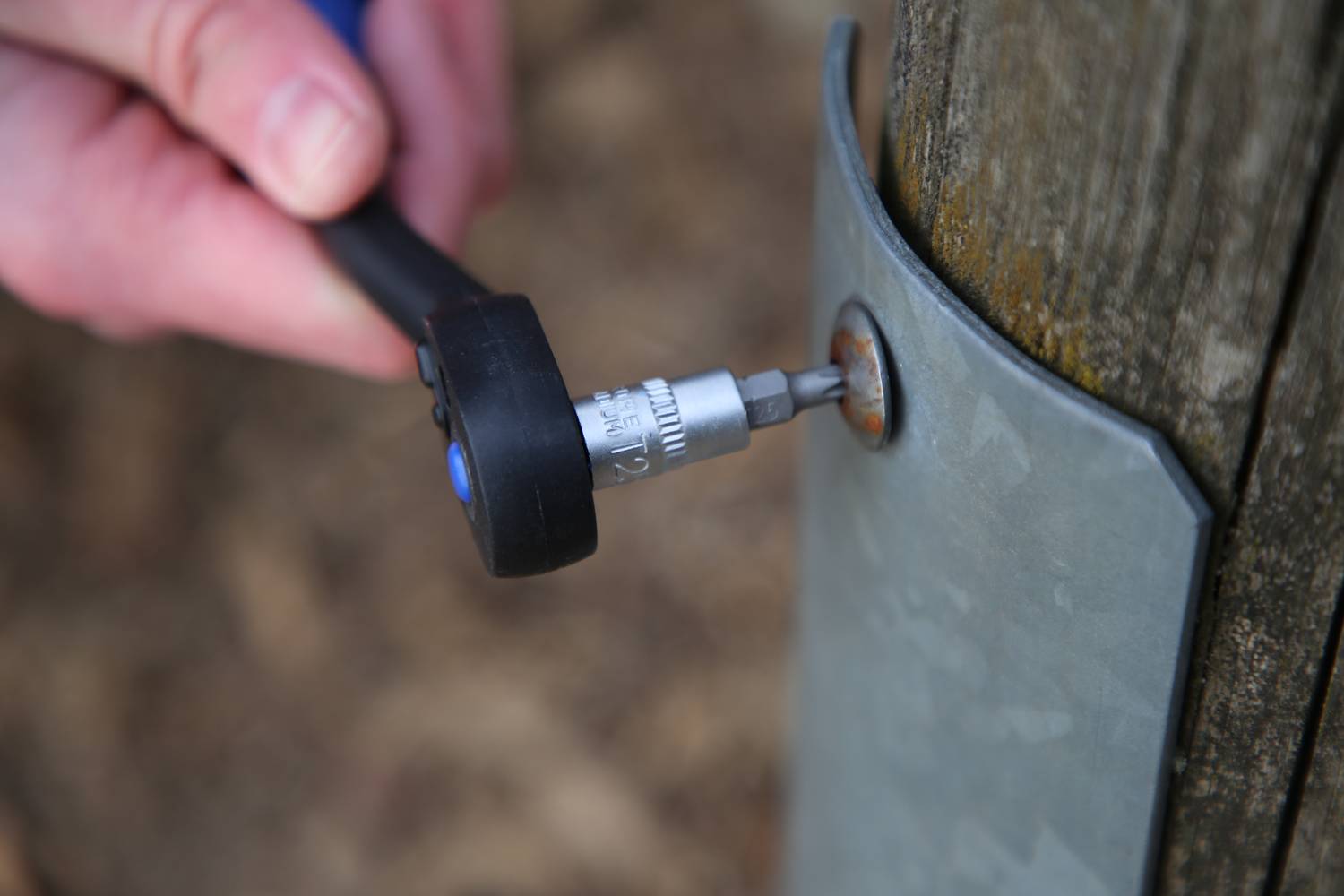 Ręka dokręca śrubę kluczem na arkuszu metalowym przymocowanym do drewnianego słupa.