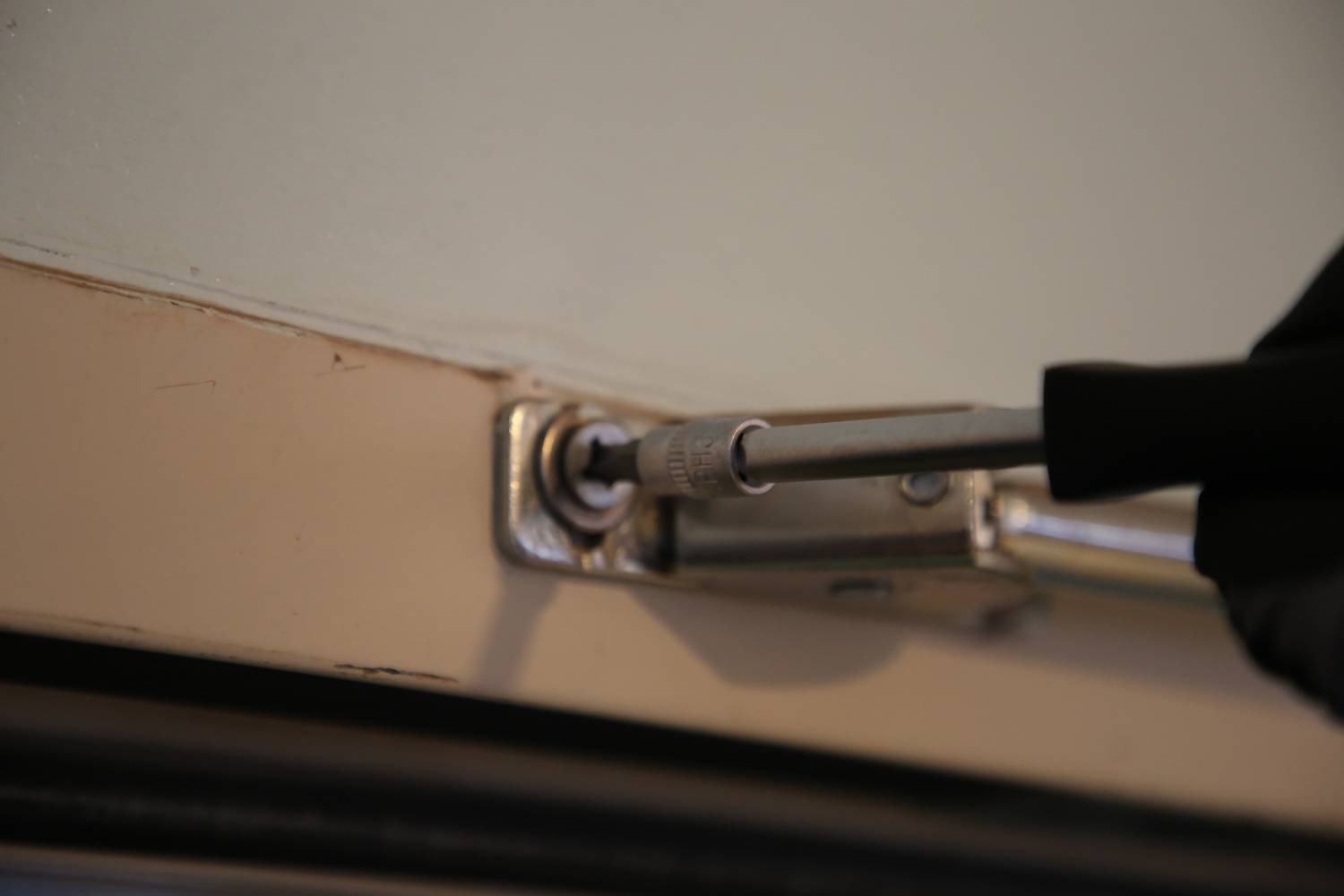 Dłoń w rękawicy dokręca śrubę w zawiasie drzwiowym, aby naprawić lub wyregulować drzwi.