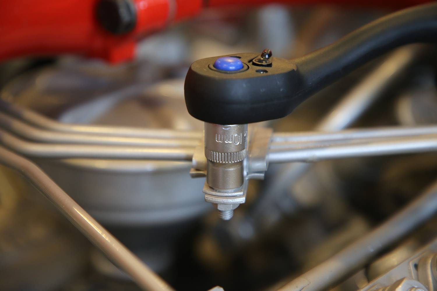 Zbliżenie klucza dynamometrycznego pracującego przy śrubie samochodu. Klucz posiada niebieski przycisk. Na rozmytym tle widoczne są części silnika.