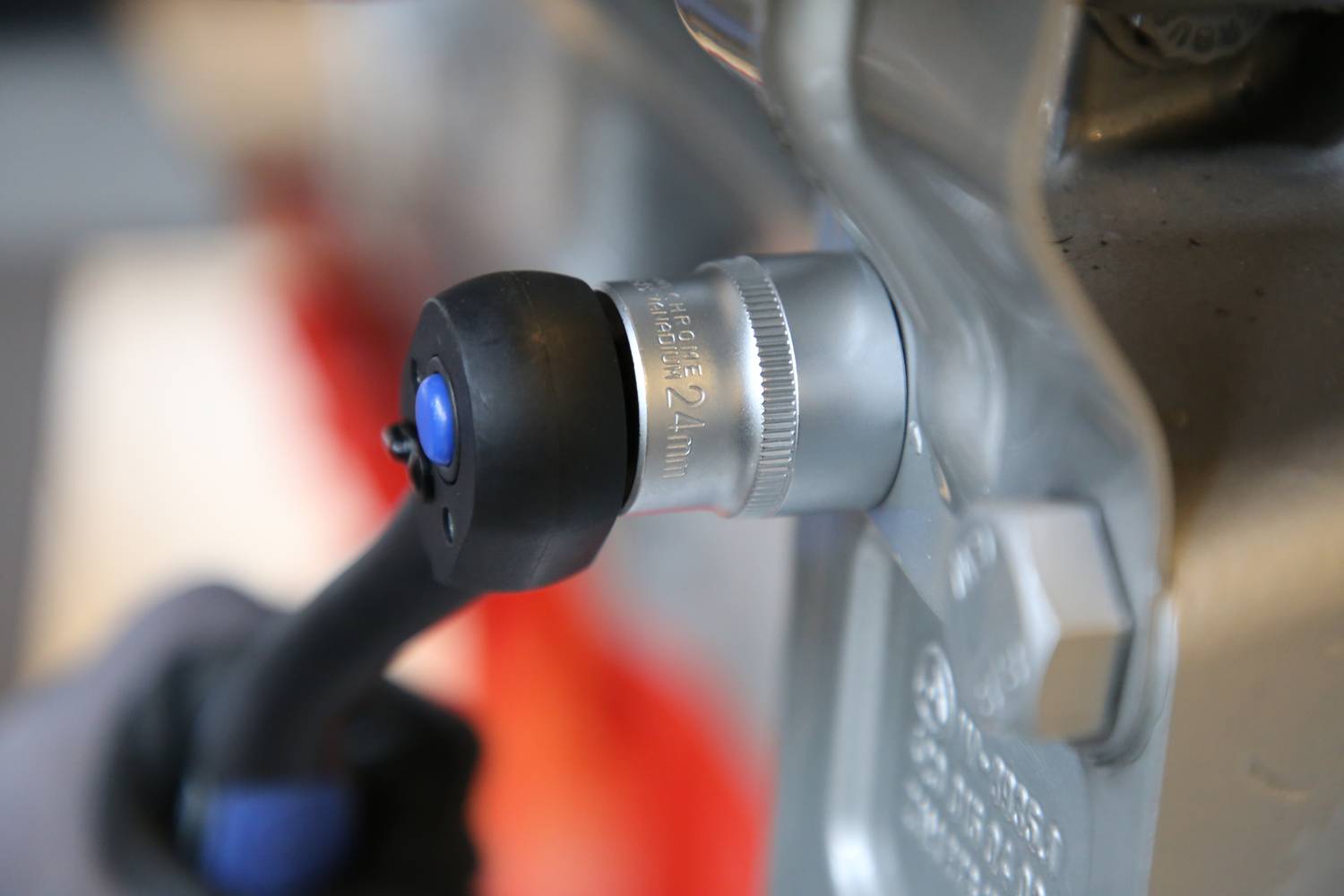 Osoba trzyma pneumatyczny klucz udarowy, celując w sześciokątną nakrętkę silnika pojazdu, aby ją odkręcić.