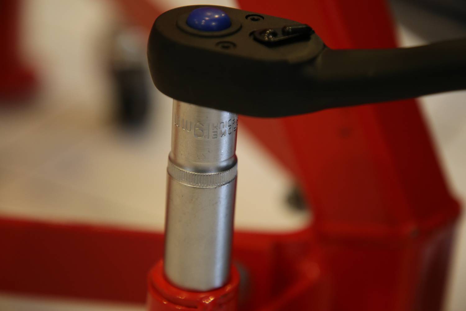 Zbliżenie metalowej dźwigni z niebieskim przyciskiem, przymocowanej do czerwonego urządzenia, prawdopodobnie będącej częścią hydraulicznego podnośnika samochodowego.