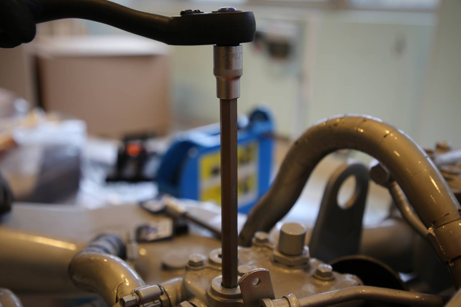 Osoba używa narzędzia do dokręcenia śruby przy silniku samochodu. W tle widać nieostre obiekty warsztatu.