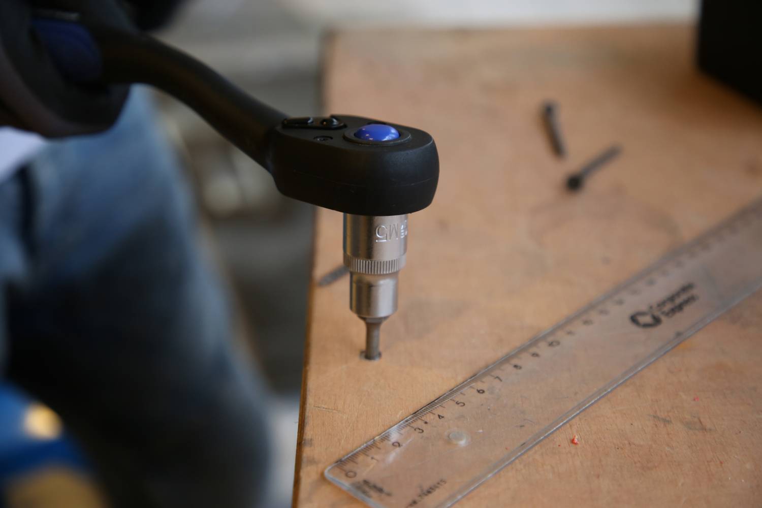 Osoba wierci otwór w desce drewnianej za pomocą wiertarki elektrycznej. Obok leży przezroczysty linijka z podziałką.