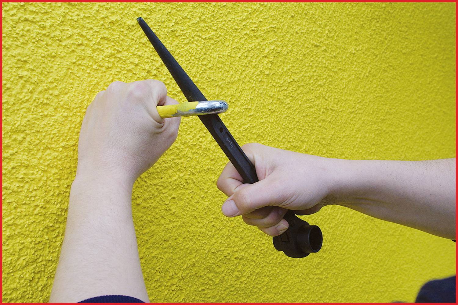 Dwie ręce trzymają narzędzia przy żółtej ścianie, jakby miały coś zamocować. Jedna ręka dzierży młotek, druga klucz hydrauliczny.