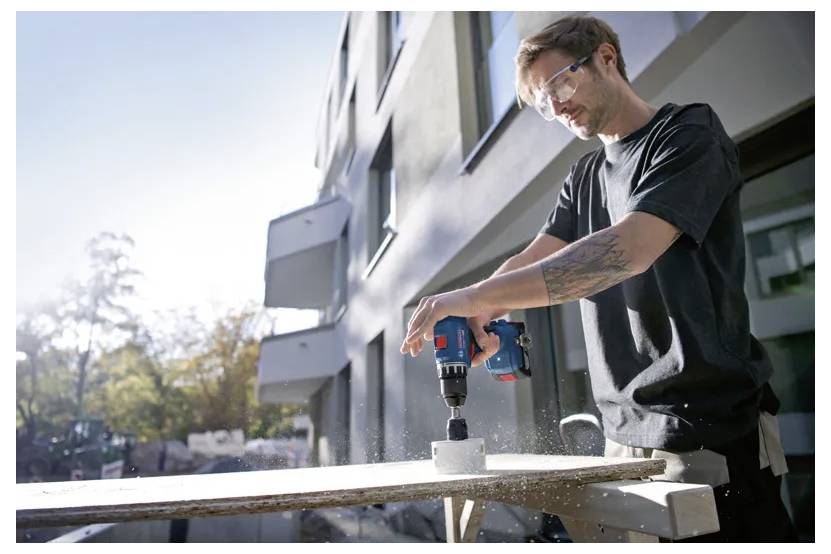 Osoba używa wiertarki elektrycznej do wykonania otworu w bloku betonowym, nosząc okulary ochronne, na zewnątrz nowoczesnego budynku.