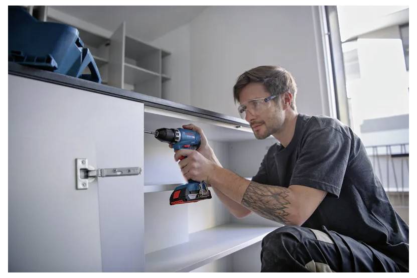 Osoba używa wiertarki elektrycznej do złożenia białej szafki, nosząc okulary ochronne i koncentrując się na mocowaniu zawiasów wewnątrz szafki.