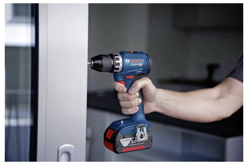 Osoba trzyma niebiesko-czarny akumulatorowy wiertarka w pobliżu metalowej powierzchni wewnątrz nowoczesnego pomieszczenia, sugerując prace majsterkowicza lub remont domu.