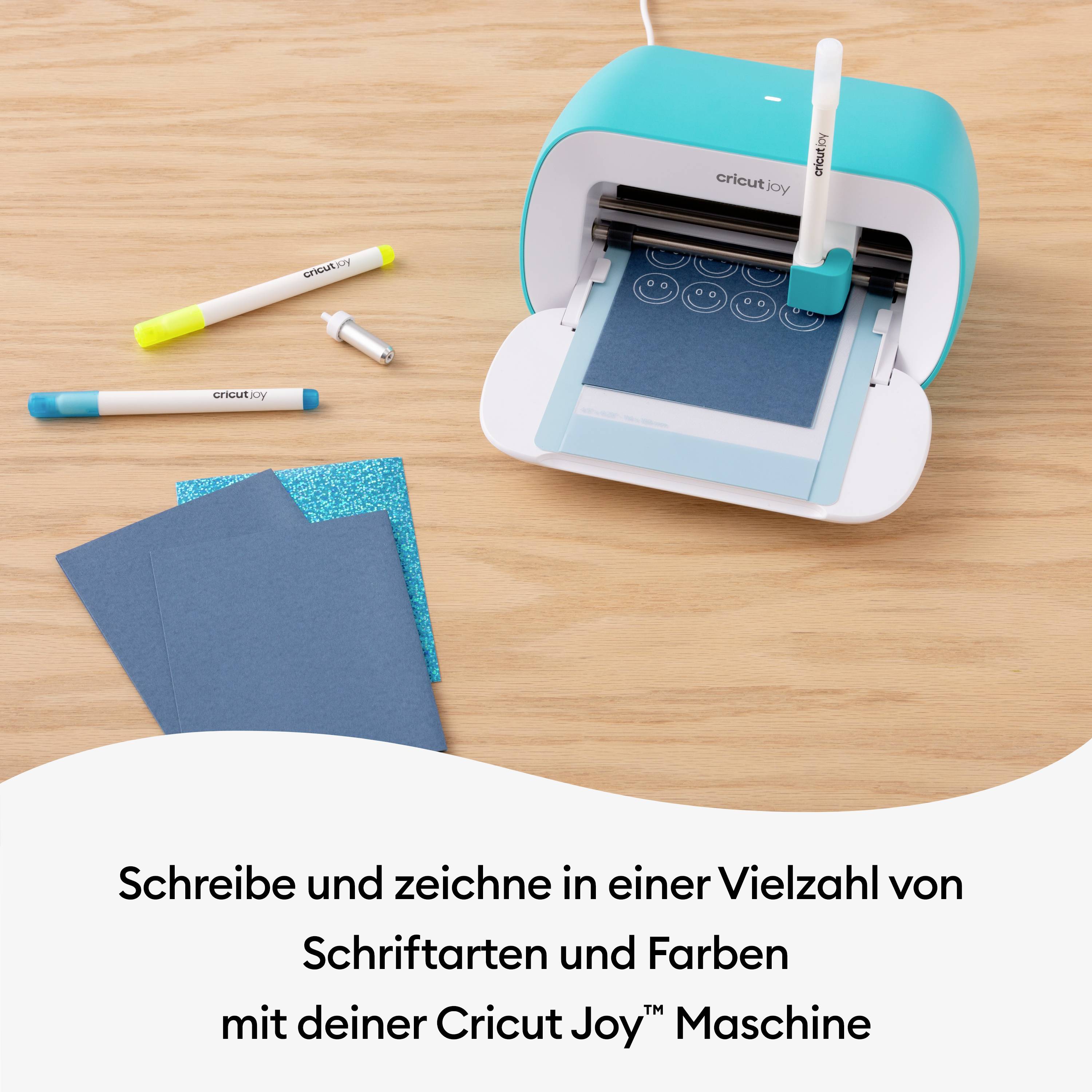 Maszyna Cricut Joy wycina papier; obok leżą pisaki i papier z brokatem. Tekst: 'Pisz i rysuj różnymi czcionkami.'
