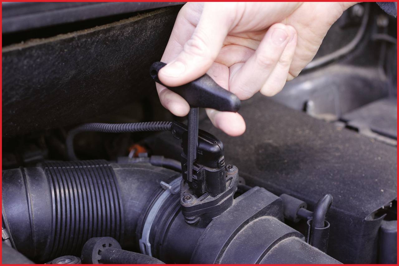 Ręka wyciąga bagnet oleju z komory silnika samochodu, aby sprawdzić poziom oleju. Silnik wykazuje typowe ślady zanieczyszczeń.