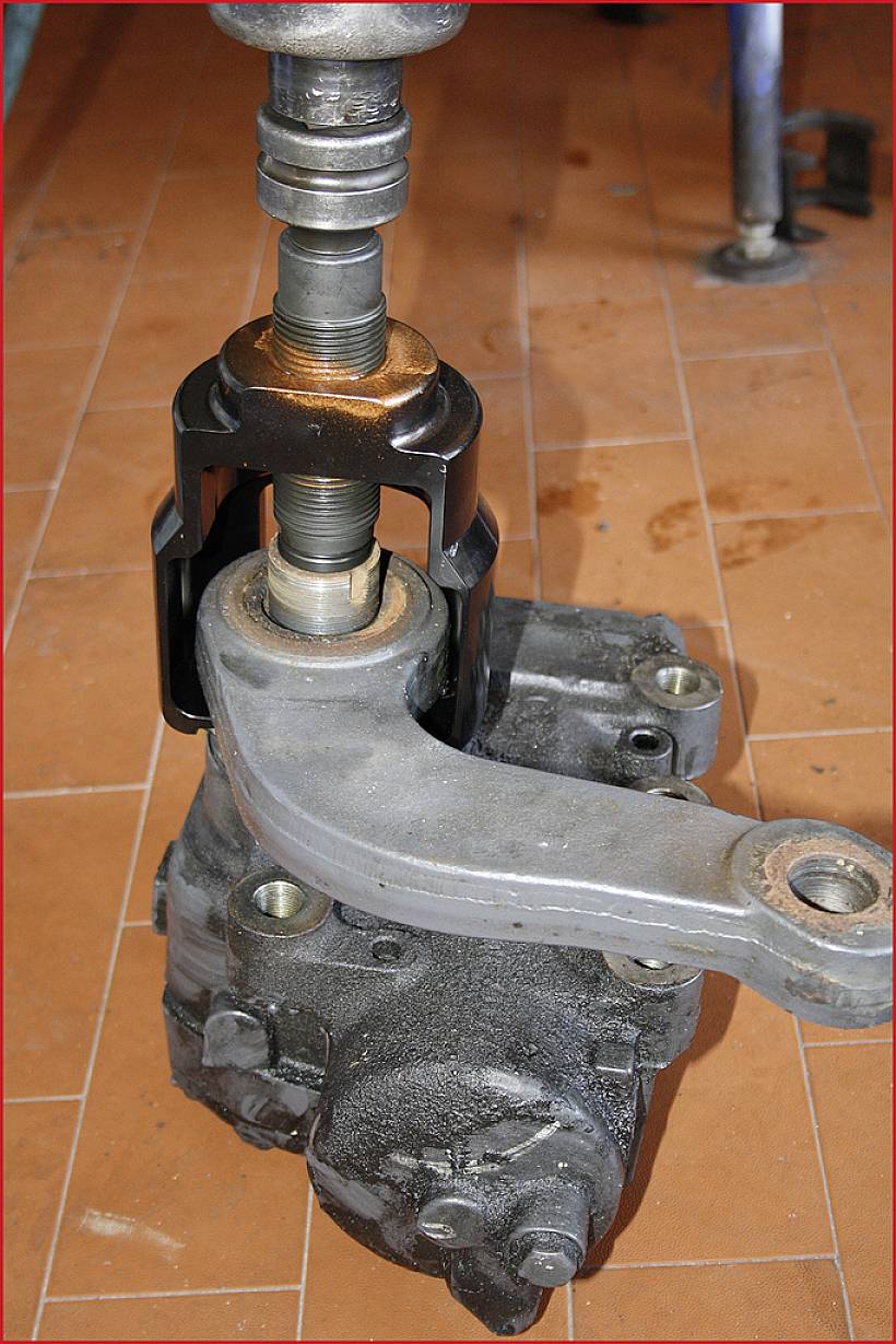 Element samochodu, prawdopodobnie wał kierowniczy z przymocowanym ramieniem dźwigni, leżący na podłodze warsztatu. Górna część obraca się mechanicznie.