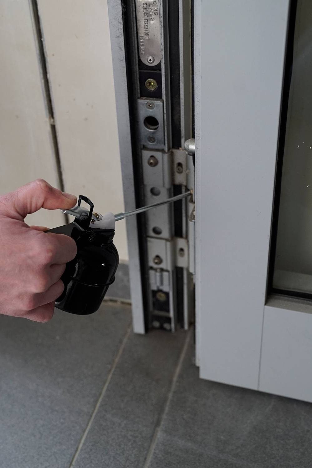 Jedna ręka trzyma narzędzie, które jest wprowadzane w ościeżnicę drzwi, aby otworzyć zamek. Podłoga wykonana jest z szarych płytek.