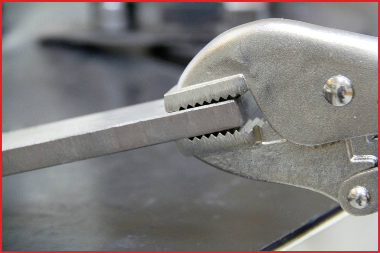 Zbliżenie szczypiec metalowych trzymających płaski kawałek metalu. Zdjęcie ukazuje szczęki szczypiec oraz rowkowany wzór uchwytu.