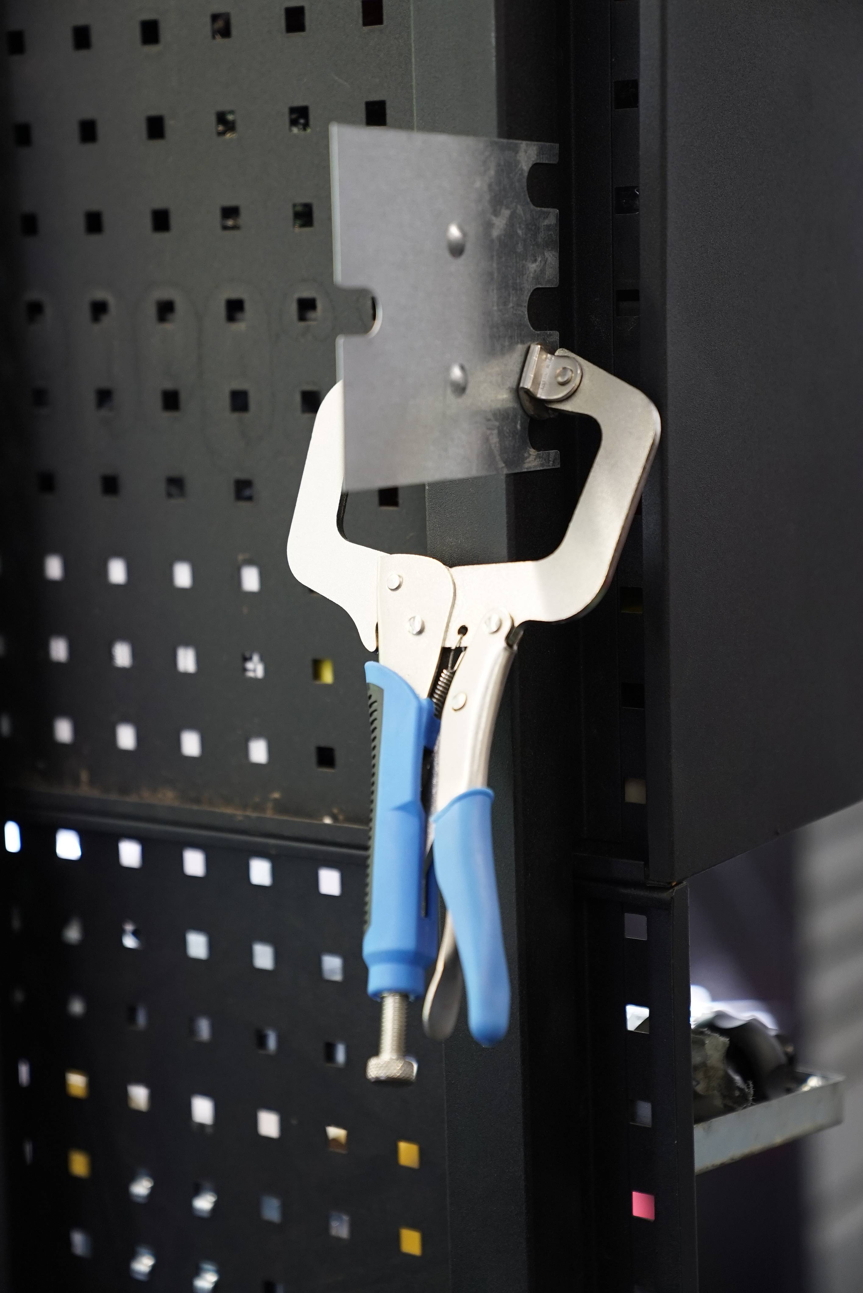 Metalowa zaciskarka z niebieskimi uchwytami przytrzymuje kawałek metalu na perforowanej blasze. Odpowiednia do prac warsztatowych lub naprawczych.