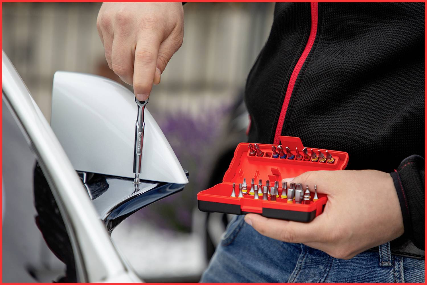 Osoba trzyma śrubokręt i zestaw końcówek śrubokrętowych w ręku, podczas gdy naprawia zewnętrzne lusterko samochodu.