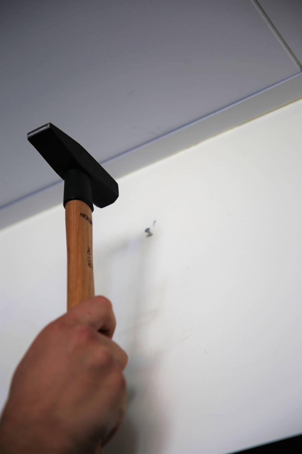 Dłoń trzyma młotek, aby wbić gwóźdź w białą ścianę.