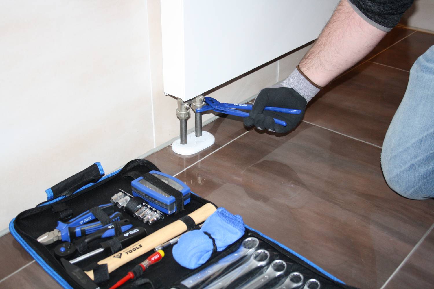 Osoba naprawia ogrzewanie kluczem hydraulicznym; skrzynka narzędziowa z narzędziami leży obok na podłodze.
