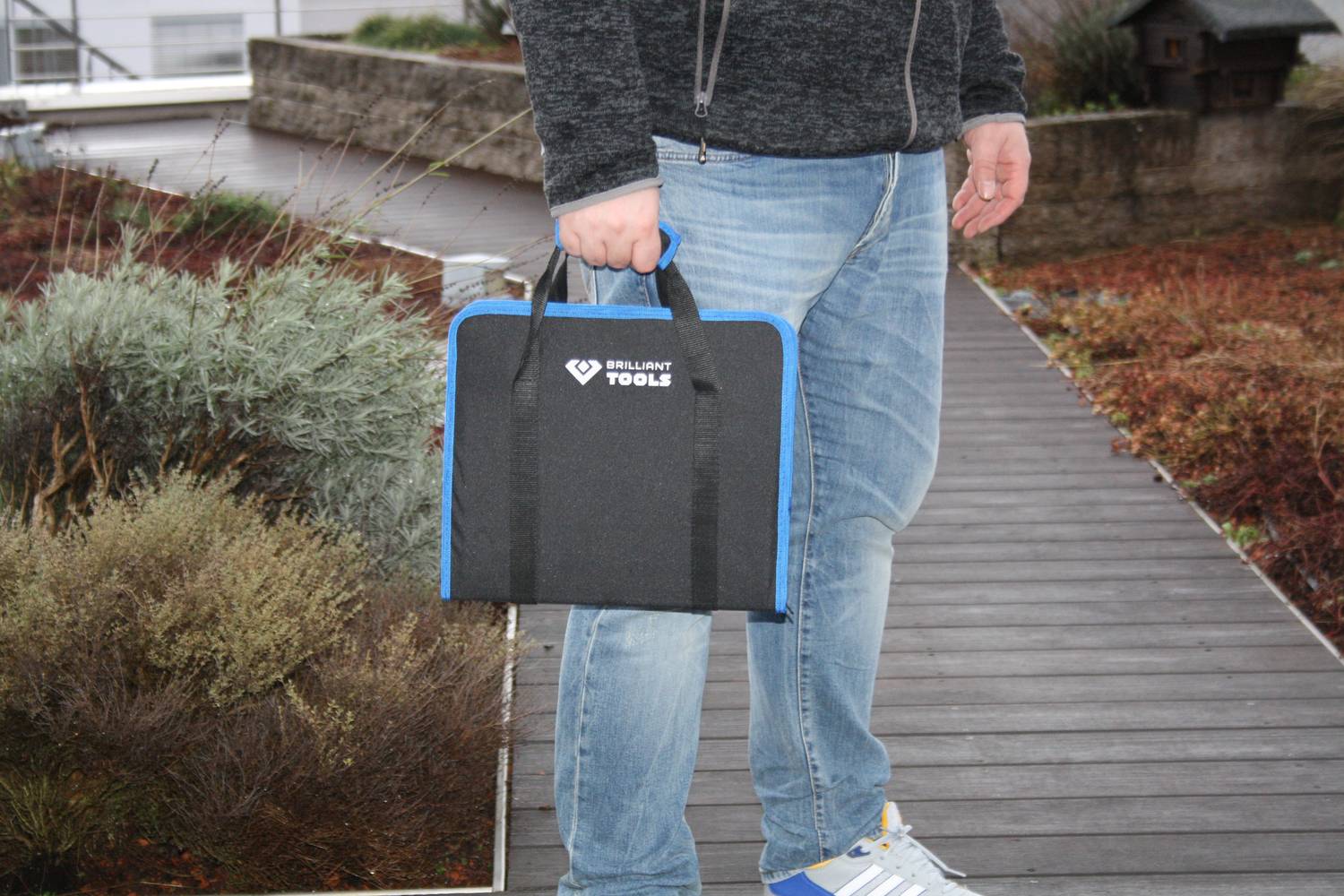 Osoba niesie walizkę z narzędziami na pomoście. Otoczenie z roślinami i budynkami.