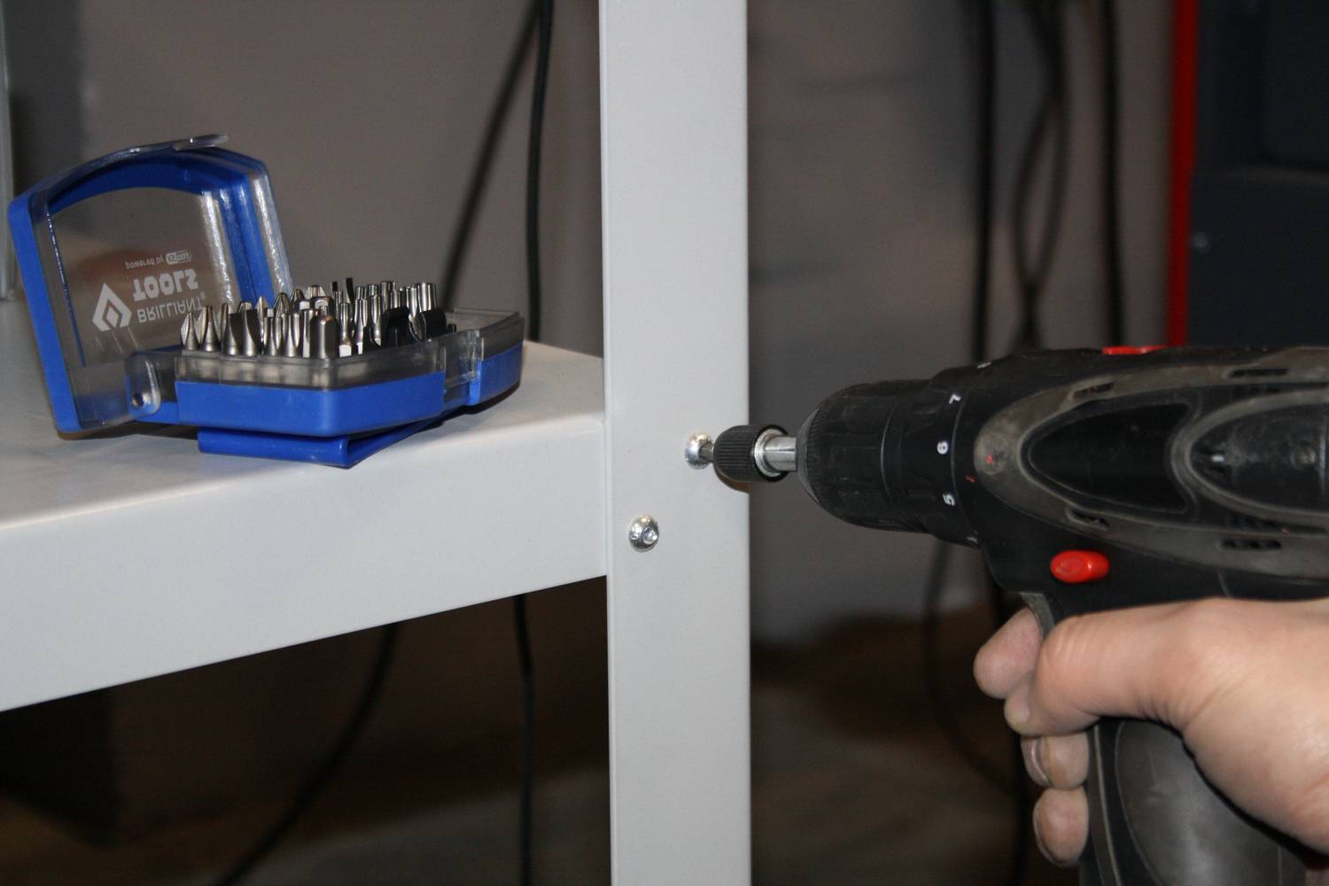 Osoba mocuje regał metalowy za pomocą wkrętarki akumulatorowej. W tle leży zestaw końcówek do wkrętów.