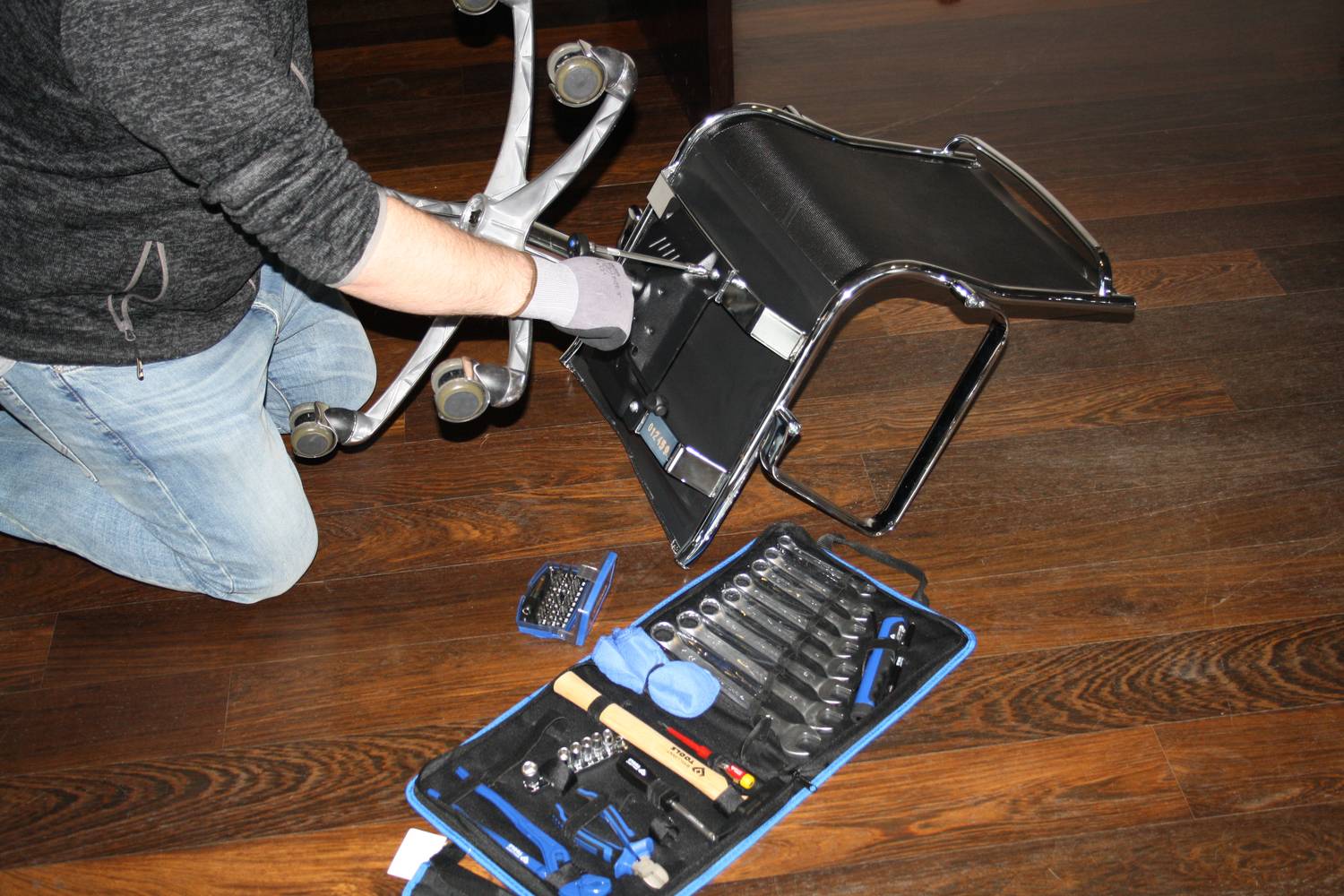 Osoba montuje fotel biurowy na drewnianej podłodze, obok której znajduje się otwarty zestaw narzędzi.