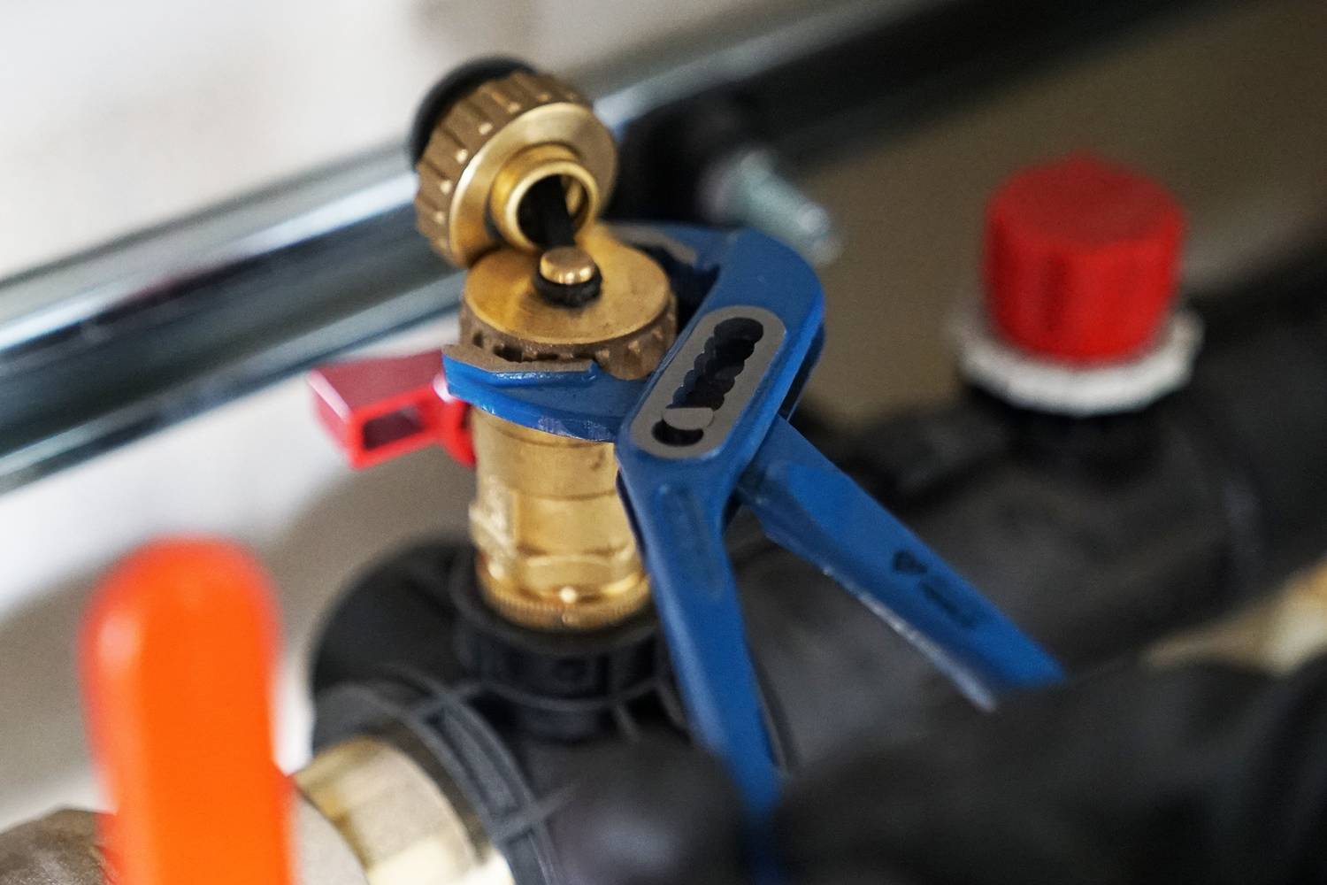 Niebieskie szczypce hydrauliczne trzymają mosiężny zawór w systemie rurowym. Zawór posiada czerwoną dźwignię. W tle widoczne są elementy instalacji rurowej.
