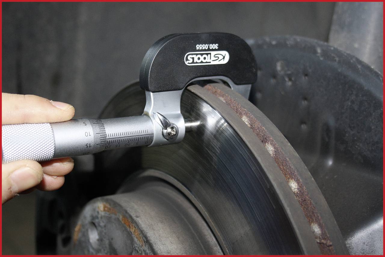 Osoba mierzy grubość tarczy hamulcowej mikrometrem. Na krawędzi tarczy widoczne są wyraźne ślady zużycia.