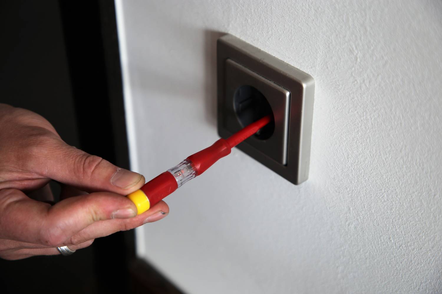 Jedna ręka trzyma tester napięcia w gnieździe ściennym. Tester jest używany do sprawdzenia napięcia elektrycznego.