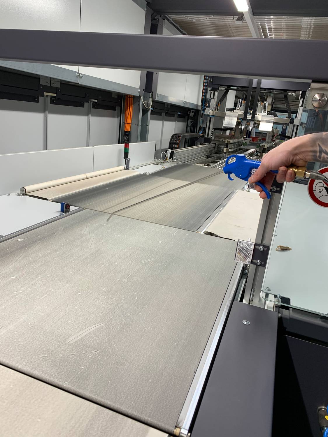 Osoba trzyma niebieskie narzędzie, pracując przy przemysłowym przenośniku, w otoczeniu maszyn na tle.