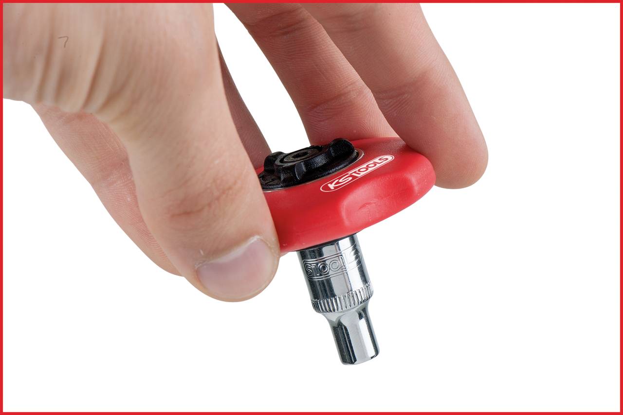 Jedna ręka trzyma czerwono-czarne narzędzie z srebrną wkładką, prawdopodobnie końcówkę klucza dynamometrycznego.