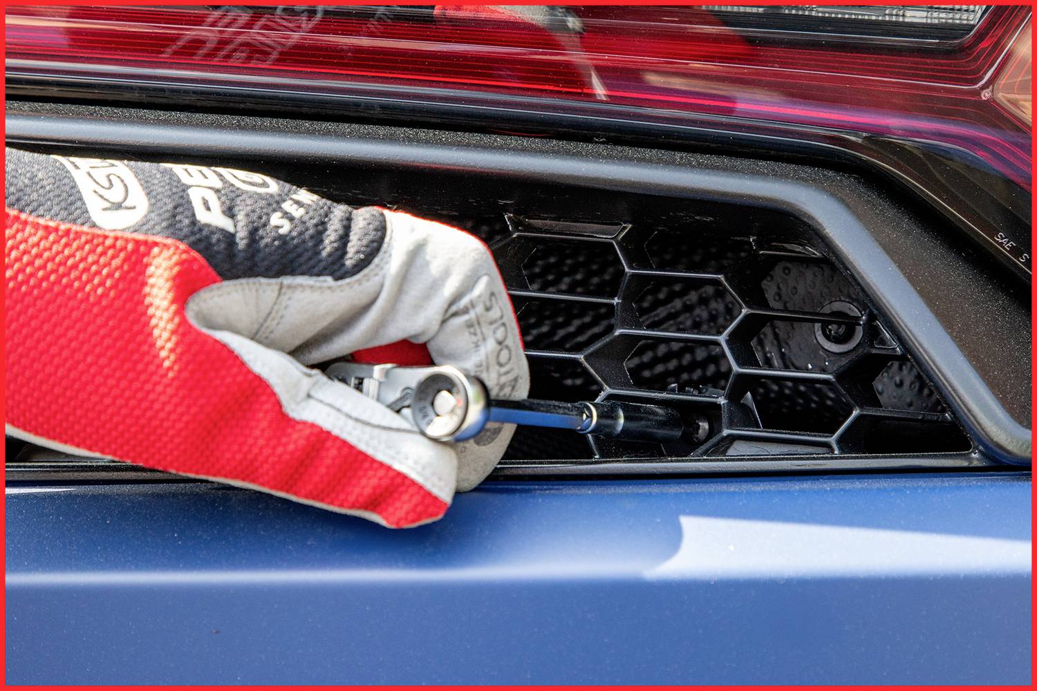 'Osoba w rękawicy używa narzędzia, aby zamocować lub zdjąć osłonę kratki plastra miodu na tylnej części pojazdu.'