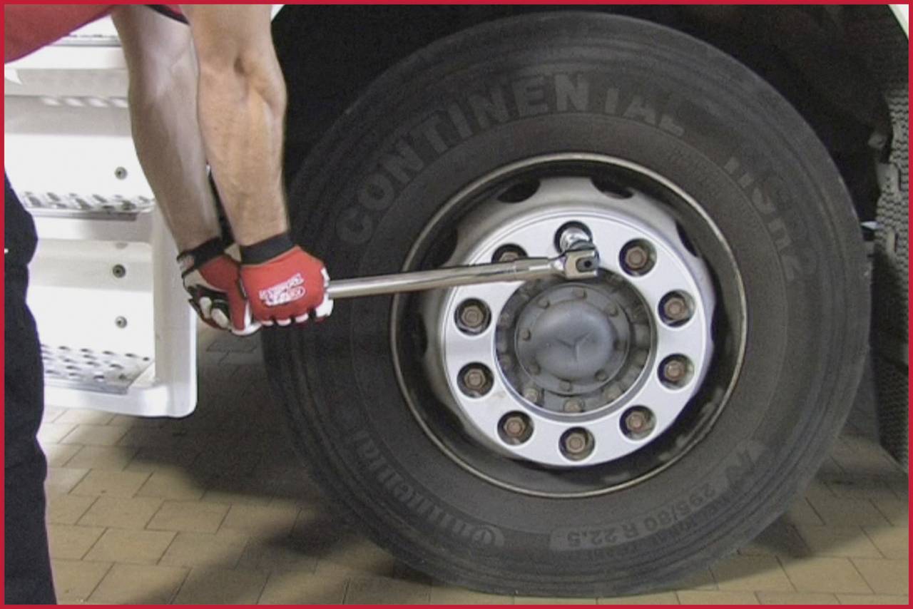 Osoba w czerwonych rękawiczkach dokręca śruby dużego koła ciężarówki kluczem nasadowym.