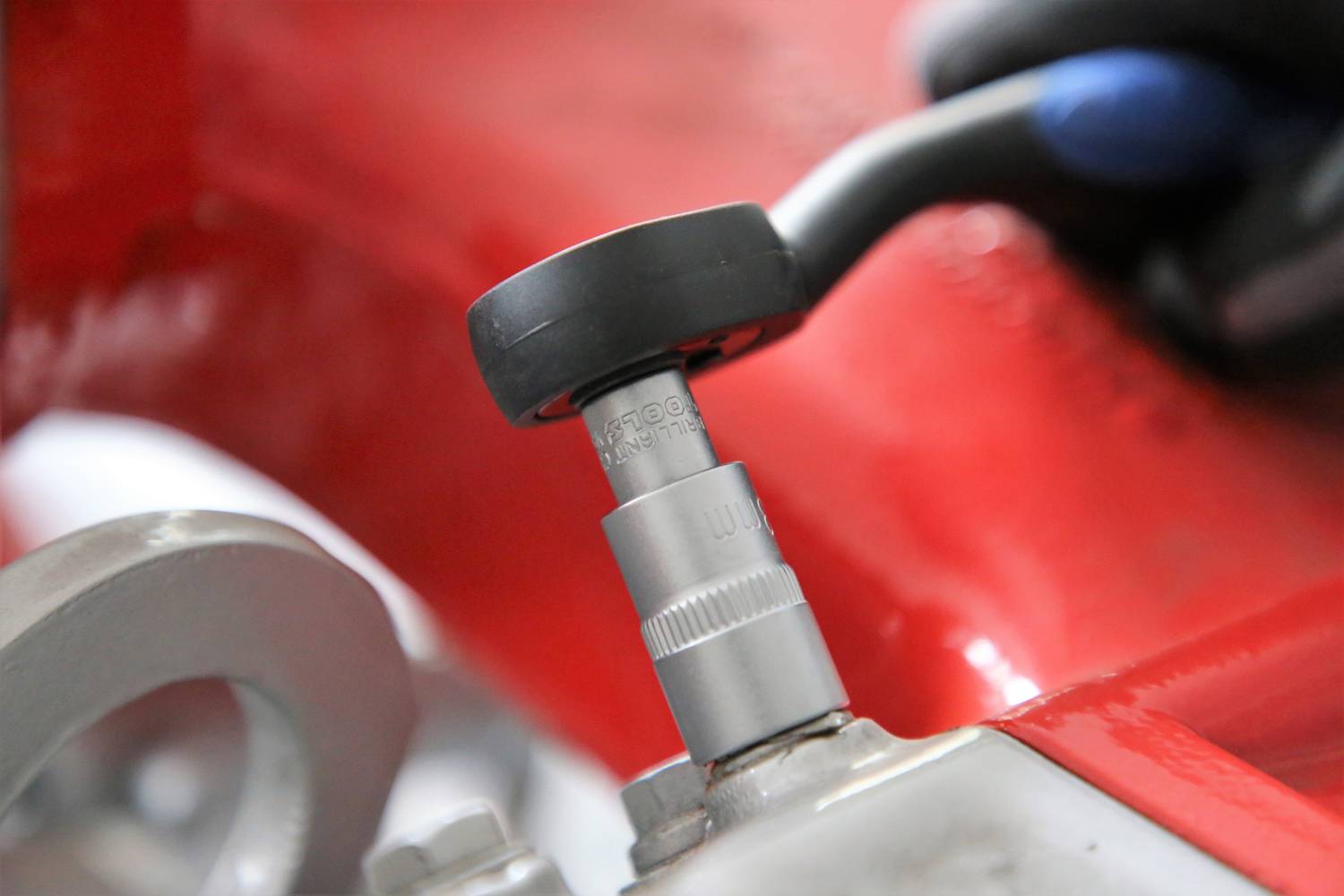 Zbliżenie klucza dokręcającego śrubę przy czerwonym elemencie maszyny. Dłoń pewnie trzyma klucz.