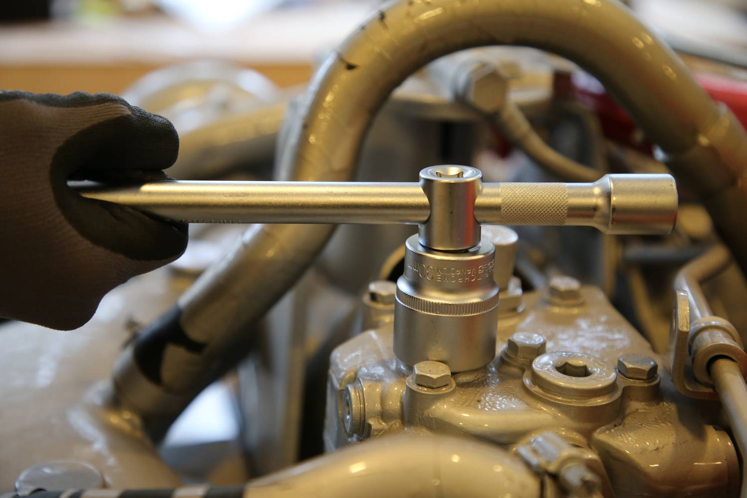 Ręka w rękawicy używa grzechotki do pracy przy maszynie. Otoczenie pokazuje przewody i części maszynowe.