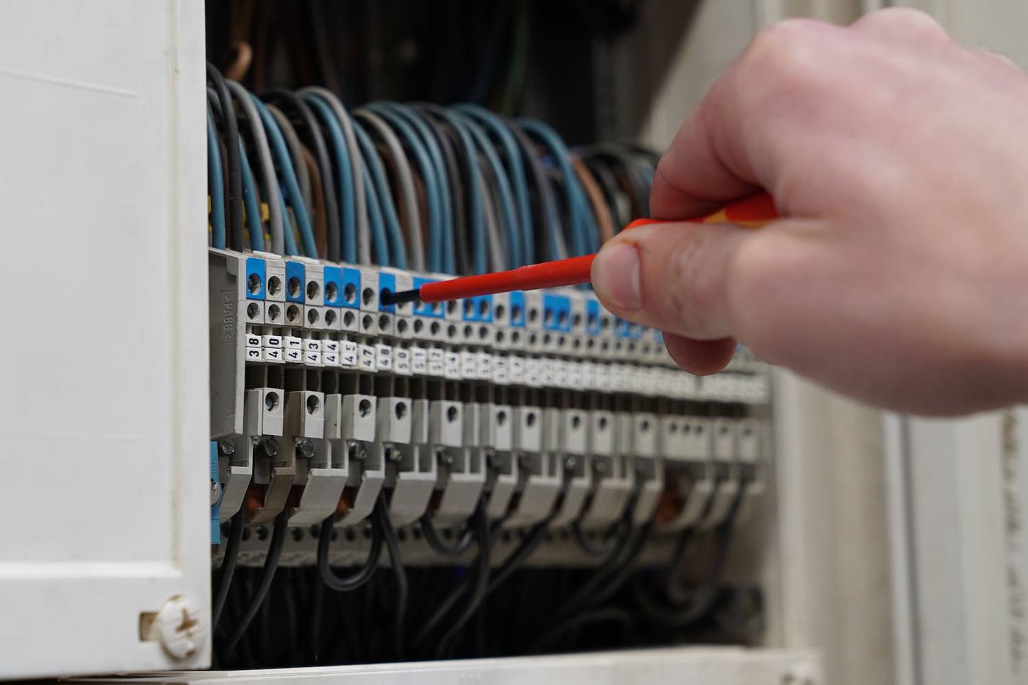 Ręka z wkrętakiem pracuje przy szafie sterowniczej z wieloma przewodami i złączami.