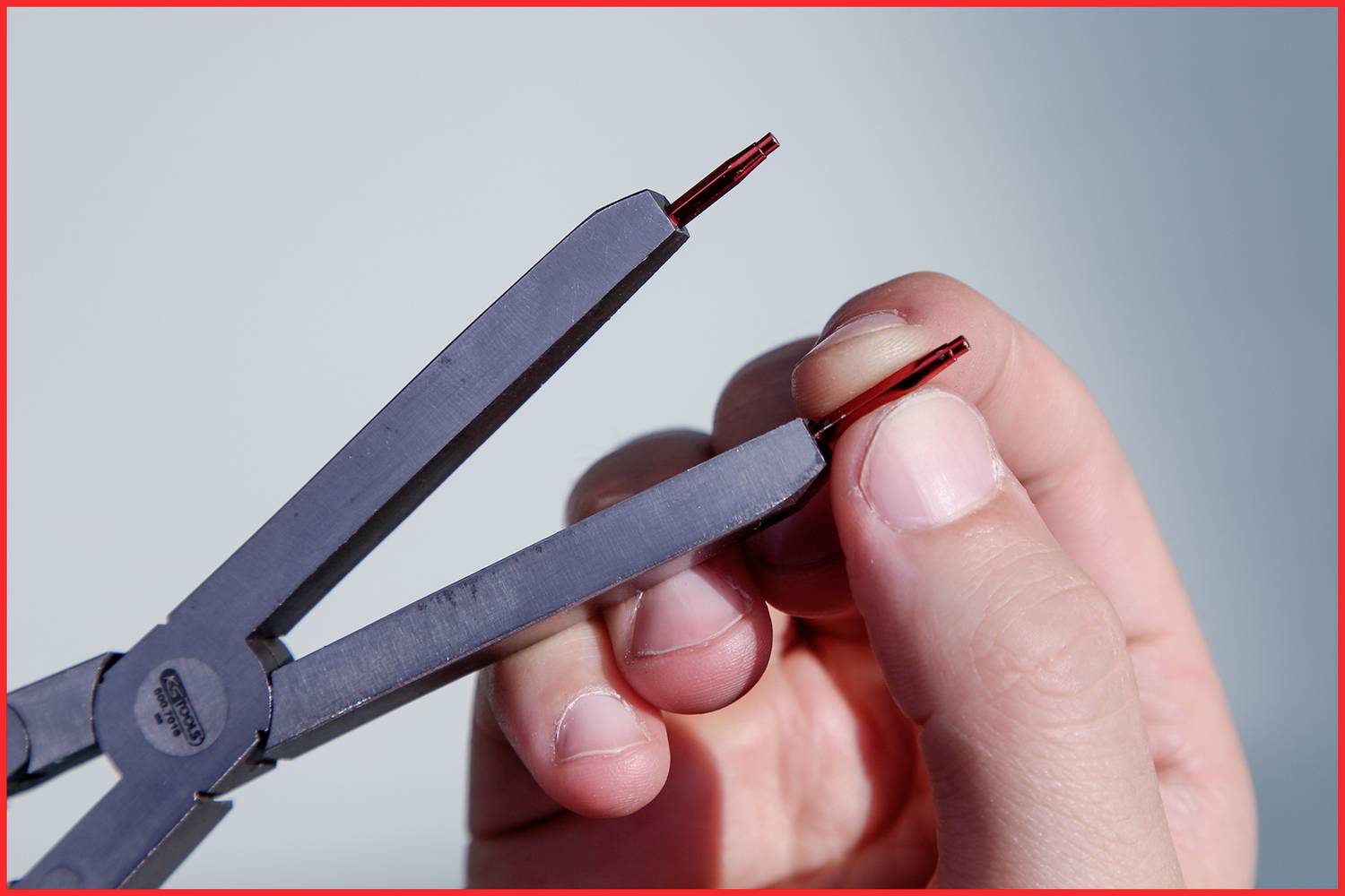 Dłoń trzyma szczypce boczne z czerwonymi przewodami między szczękami. Szczypce są podtrzymywane lewą ręką.