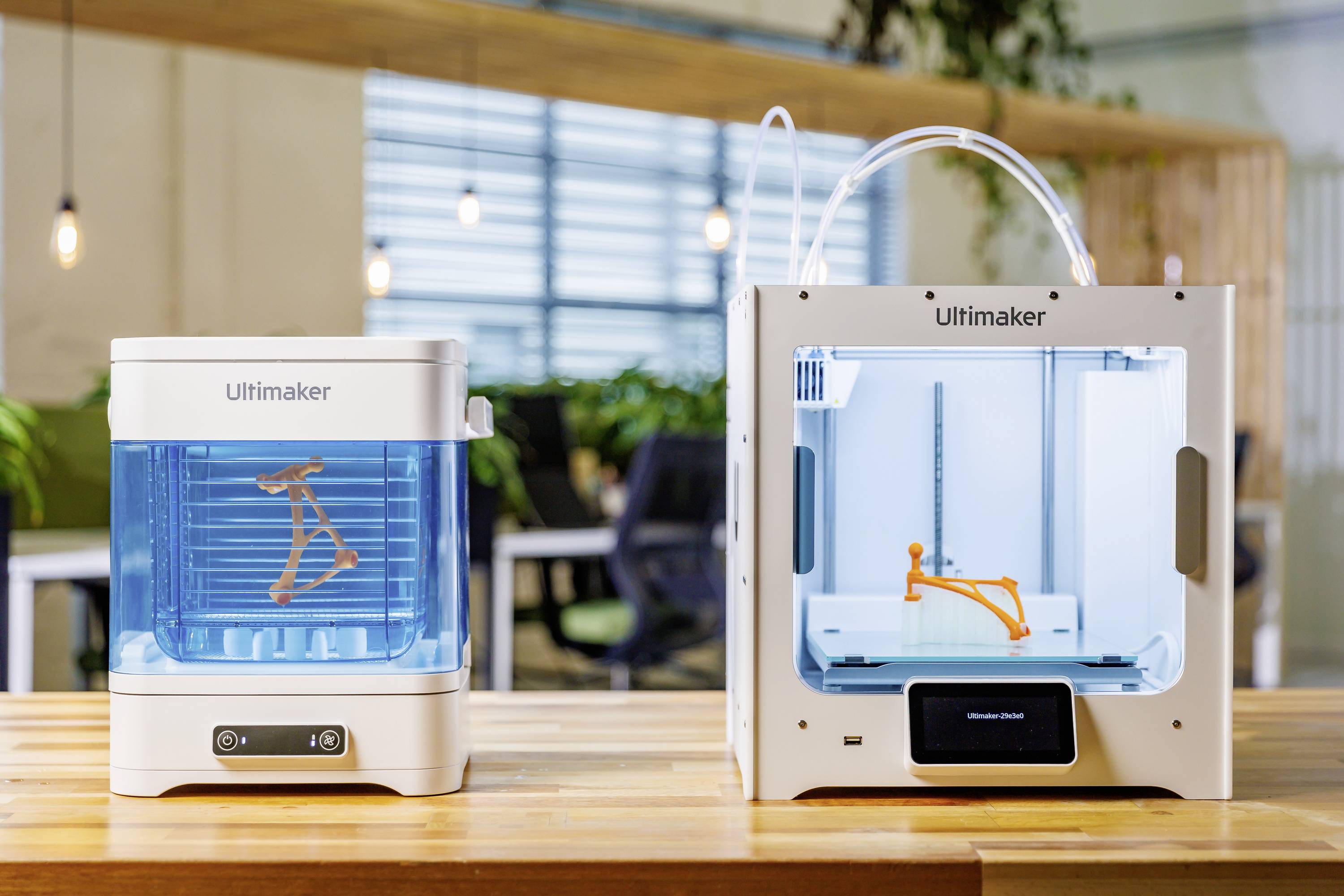 Dwa drukarki 3D Ultimaker na drewnianym stole w nowoczesnym biurze. Po lewej stronie stacja czyszcząca, po prawej drukarka z pomarańczowym modelem.