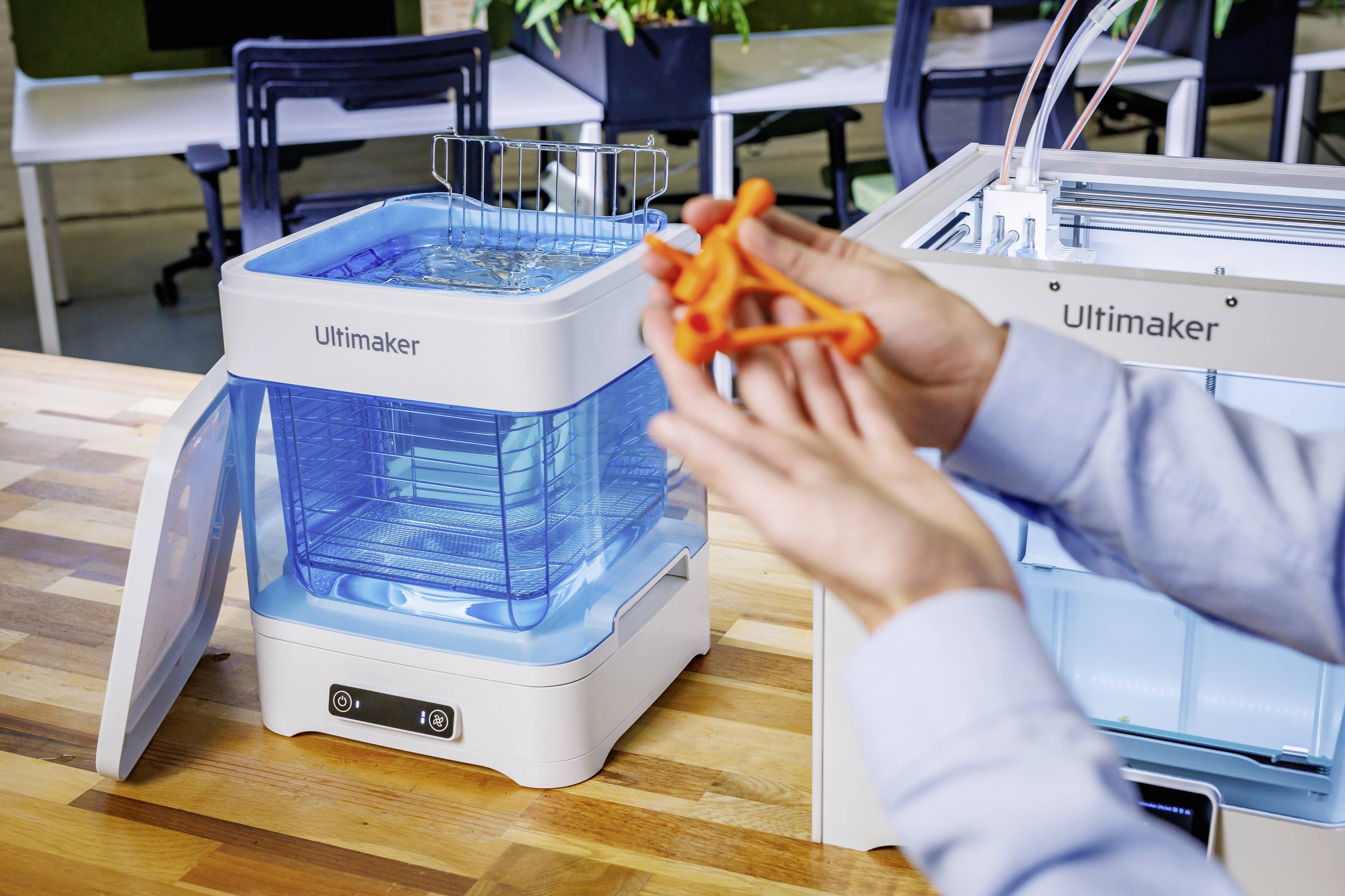 Osoba trzyma pomarańczowy obiekt wydrukowany w technologii 3D przed niebieskim urządzeniem czyszczącym firmy Ultimaker, stojącym na drewnianym stole.