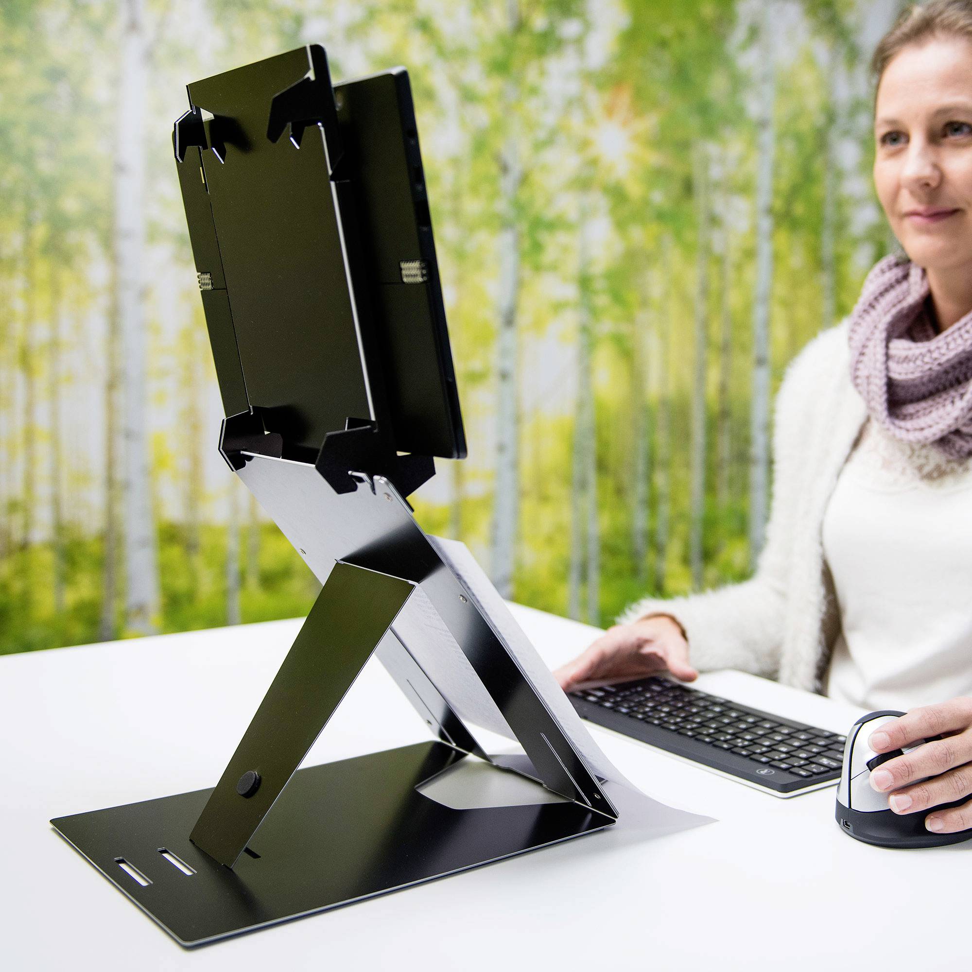 Osoba siedzi przy stole i używa urządzenia umieszczonego na stojaku, otoczona zielonym tłem drzew.