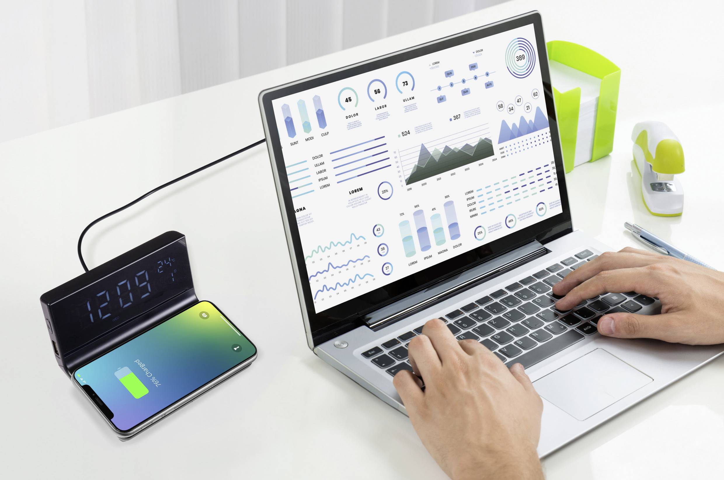 Osoba pisze na laptopie, na którego ekranie wyświetlają się wykresy danych. Obok, po prawej stronie, smartfon ładuje się bezprzewodowo, a zegar wskazuje godzinę 12:09.