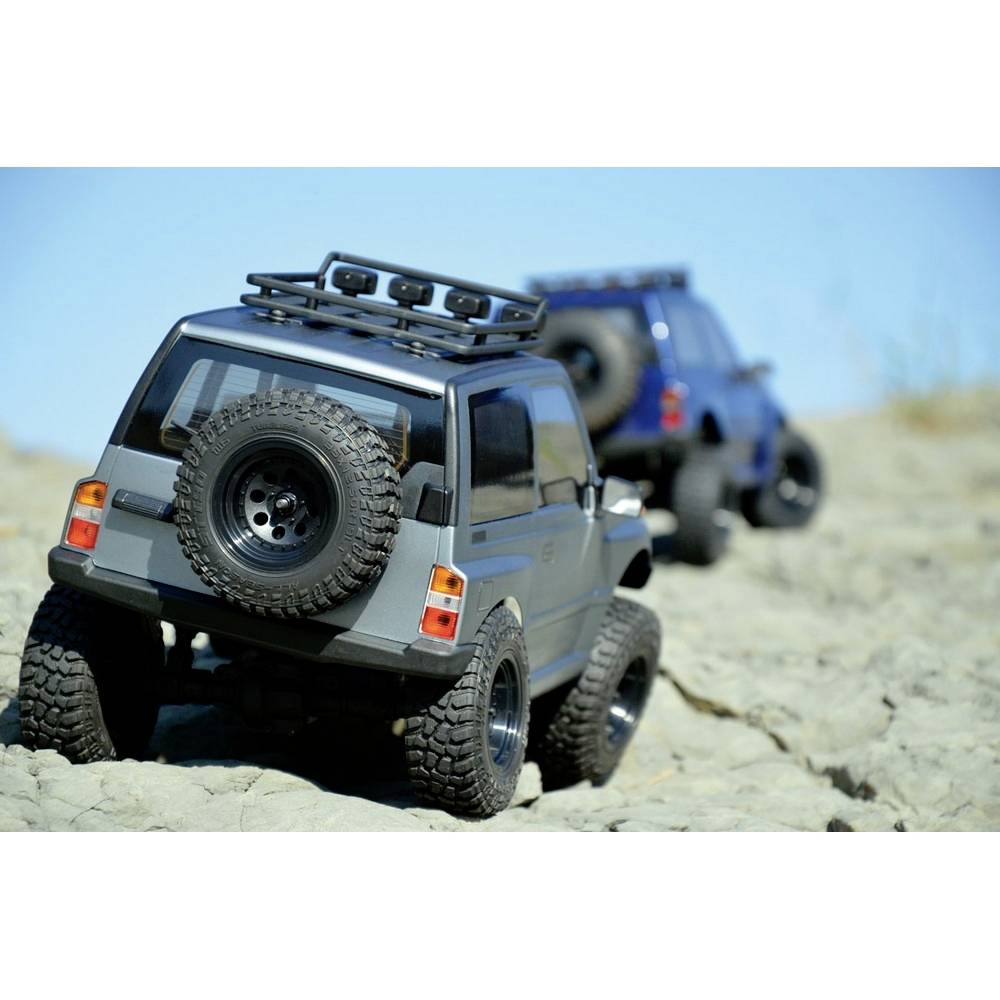 Dwa zdalnie sterowane terenówki jadą po nierównym, skalistym terenie przy bezchmurnym niebie. Focus jest skierowany na przedni, szary samochód.