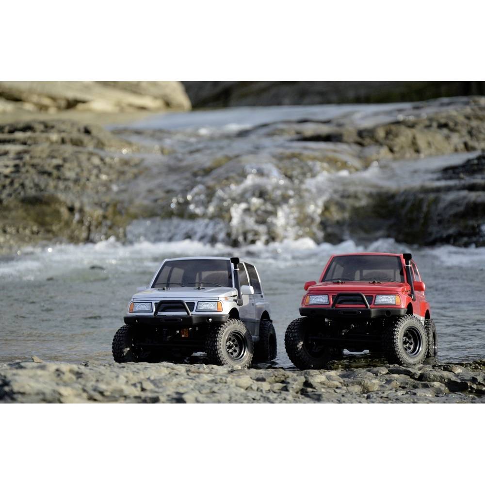 Dwa zdalnie sterowane samochodziki modelowe w kolorach czerwonym i szarym stoją na skalistym gruncie w pobliżu płynącego strumienia na wolnym powietrzu.