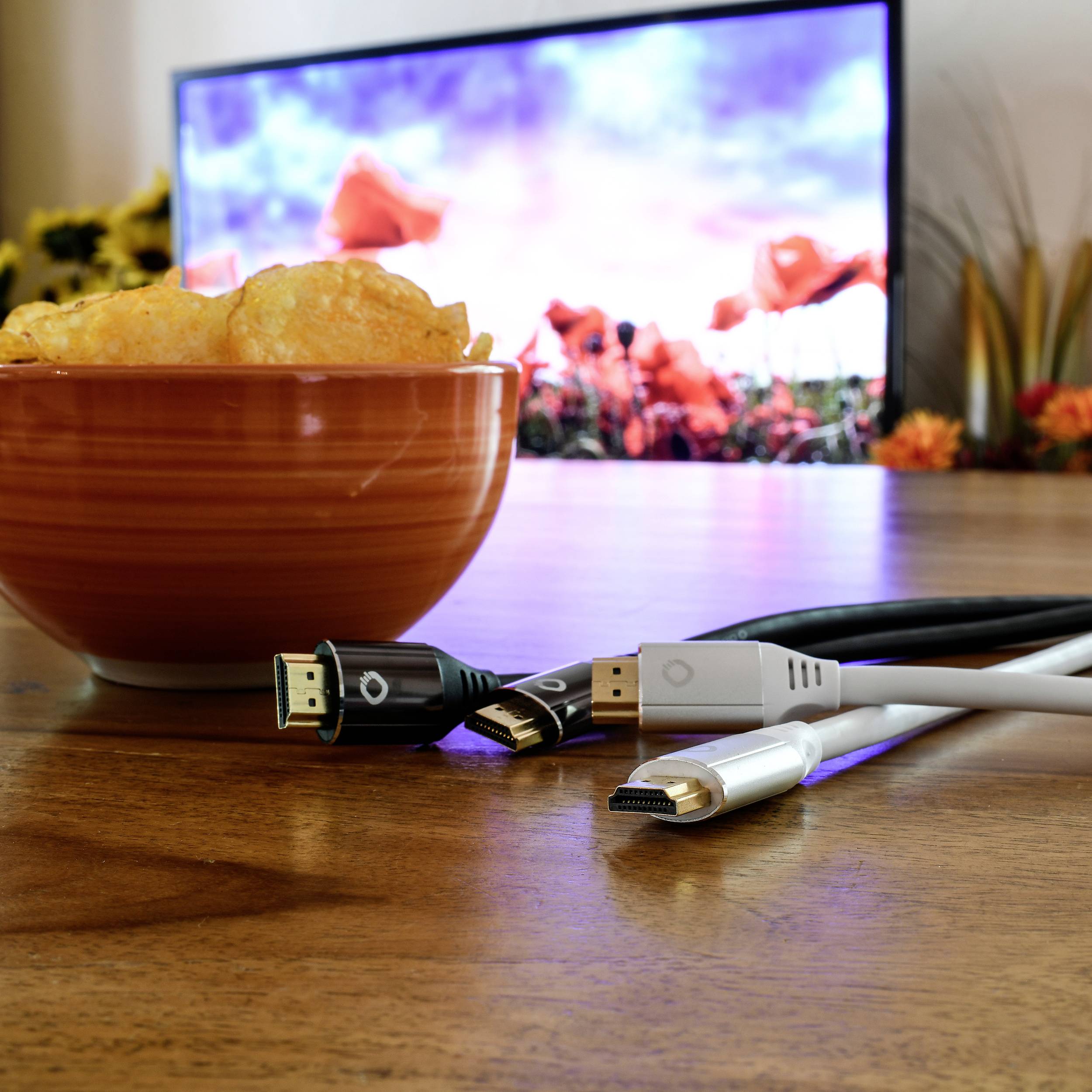 Miska z chipsami ziemniaczanymi stoi na stole przed telewizorem wyświetlającym kolorowy krajobraz kwiatowy. Obok leżą kable.