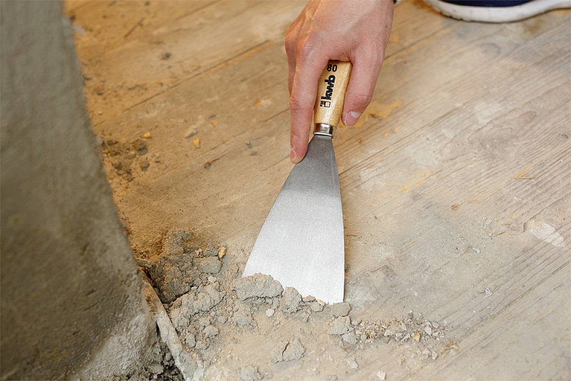 Ręka używa szpachelki do usuwania suchej zaprawy z drewnianej podłogi w pobliżu ściany.