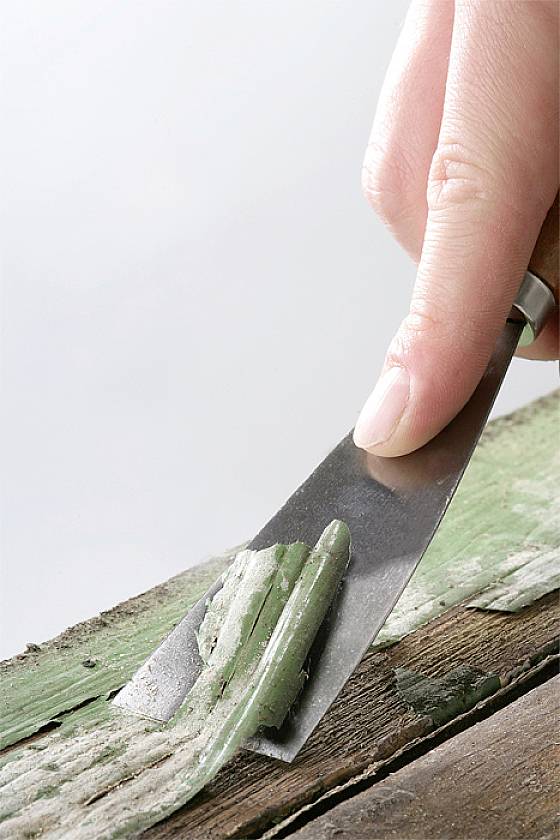 Dłoń trzyma szpachelkę i usuwa farbę ze starej powierzchni drewnianej.