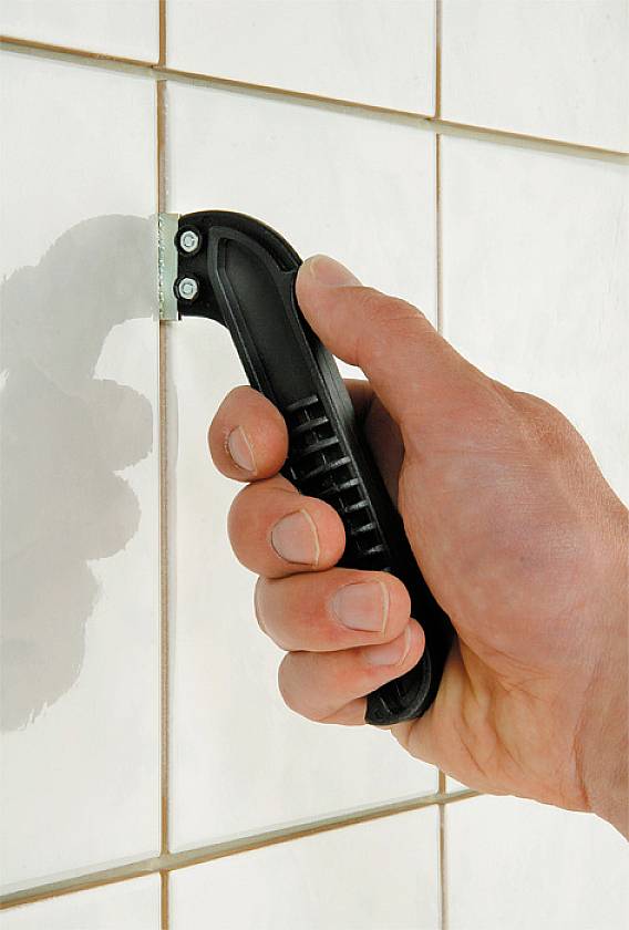 Dłoń trzyma skrobaczkę do fug i usuwa zaprawę między białymi płytkami na ścianie.
