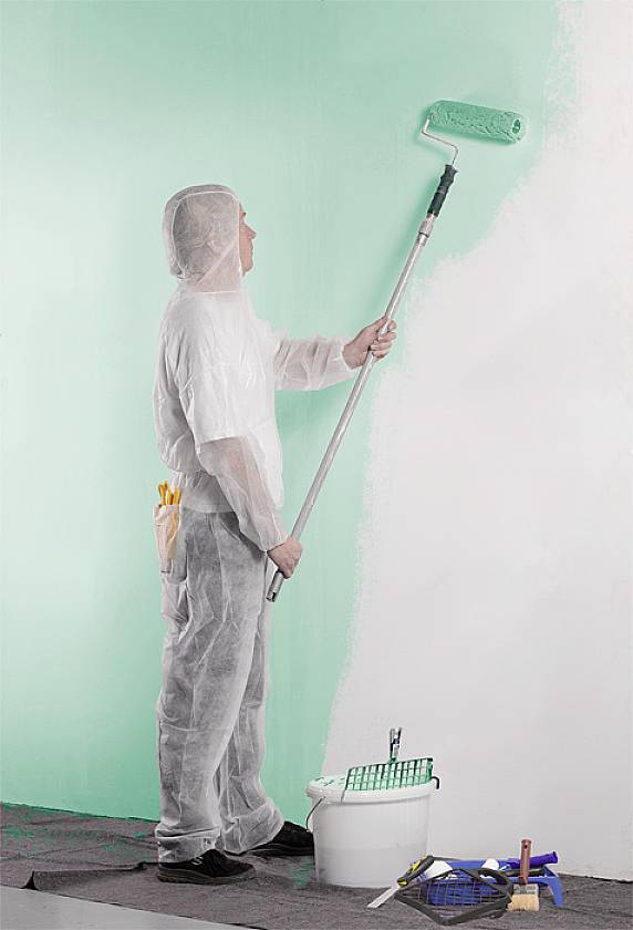 Osoba maluje ścianę na zielono wałkiem malarskim. Ma na sobie odzież ochronną i stoi obok wiadra z farbą oraz niezbędnych akcesoriów.