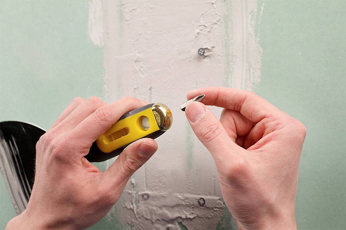 Dwie ręce trzymają żółte narzędzie i śrubę przed ścianą.Fragment ściany jest wyrychtowany tynkiem i szpachlowany.