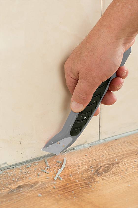 Ręka trzyma narzędzie wielofunkcyjne i usuwa materiał wypełniający spoinę między płytką ścienną a drewnianą podłogą.